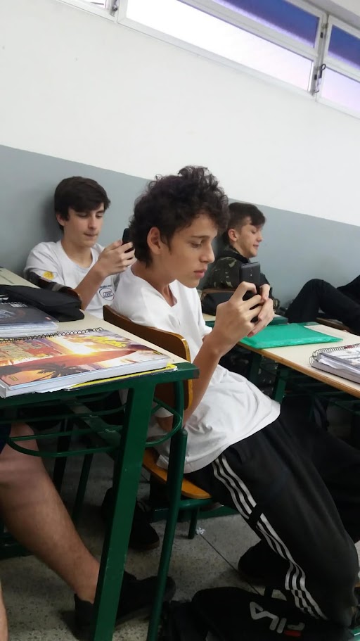 Iago adolescente na sala de aula, mexendo no celular entre colegas