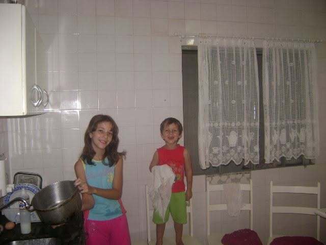 Iago e a irmã crianças na cozinha, ela na pia com uma panela e ele segurando um pano de prato