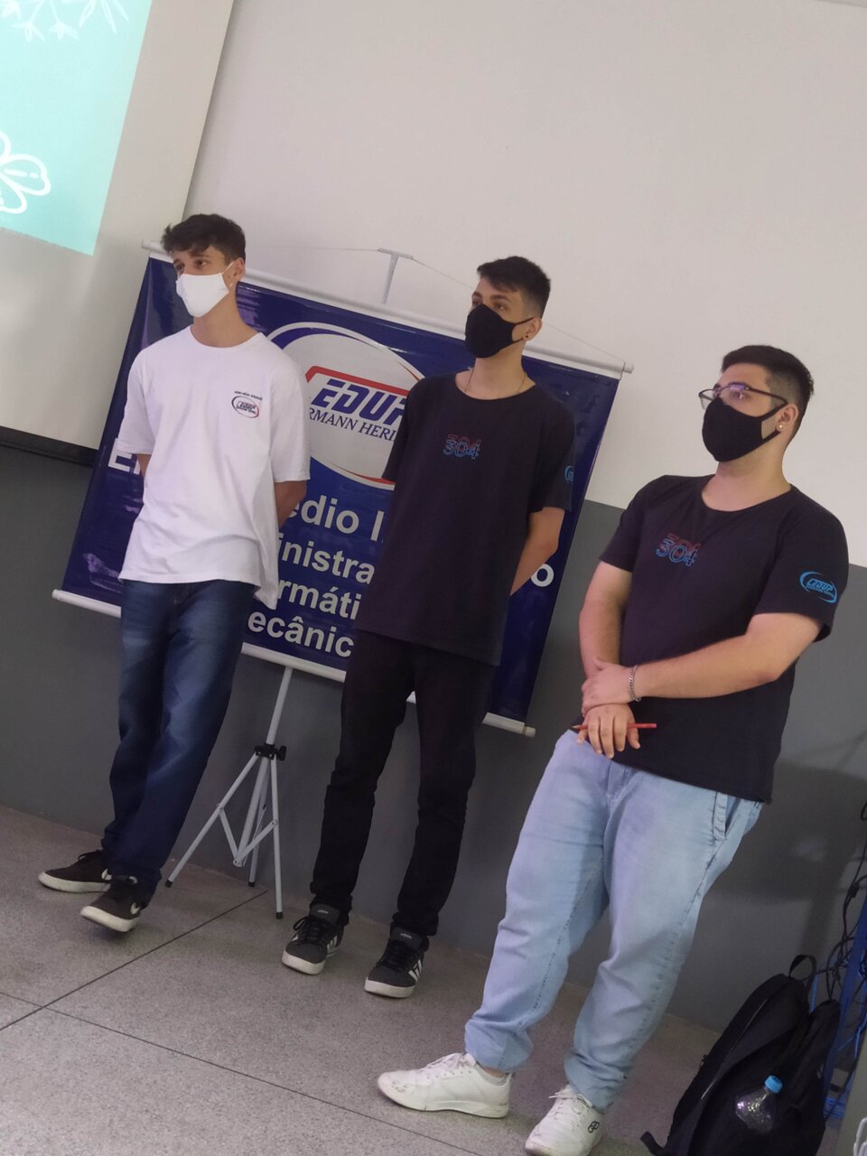 Três alunos de máscara em frente ao banner do CEDUP Hermann Hering, apresentando um trabalho